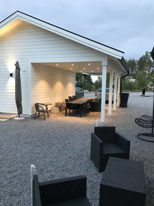 Nybyggd vit carport med inbyggd belysning och en matplats möblerad med bord och stolar.