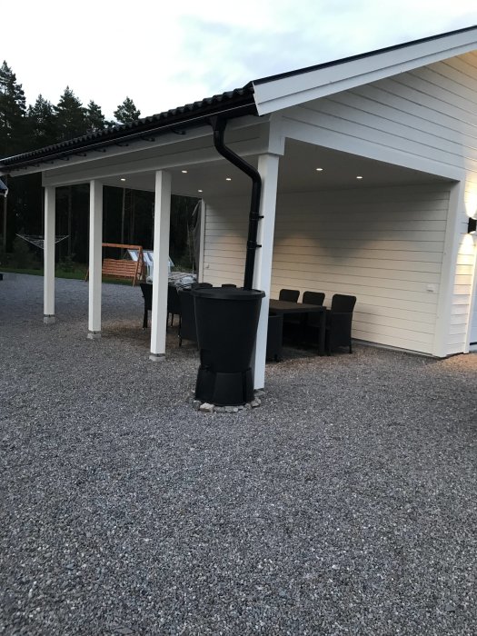 Nybyggd vit carport med svarta takrännor, belysning, uteplats med möbler och grusgång.