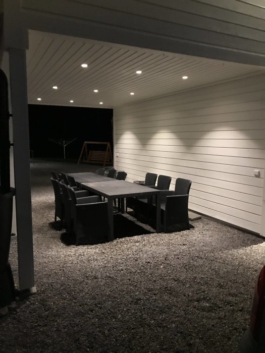 Upplyst uteplats med ett stort mörkt matbord och stolar under ett tak med inbyggda spotlights på kvällen.