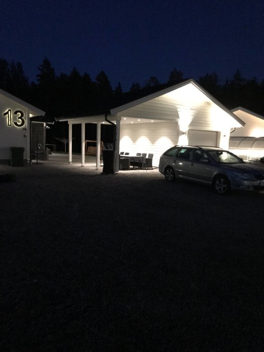 En upplyst villa och carport på kvällen med numret 13 synligt och en bil parkerad framför.