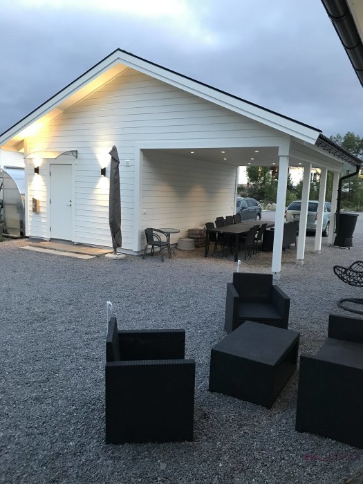 Nybyggd vit carport med inbyggd belysning och ett uteplatsområde med svarta möbler.