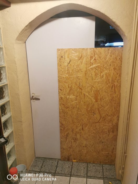 En tillfällig dörr av plywood täcker ett dörrhål mellan förråd och garage i ett hus.