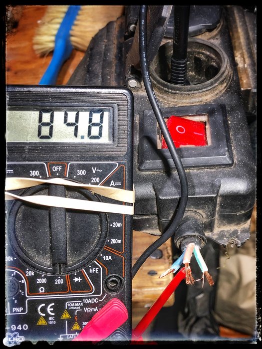 Elektrisk multimeter visar 14,8V ansluten till en klippt förlängningssladd med synliga ledningar.