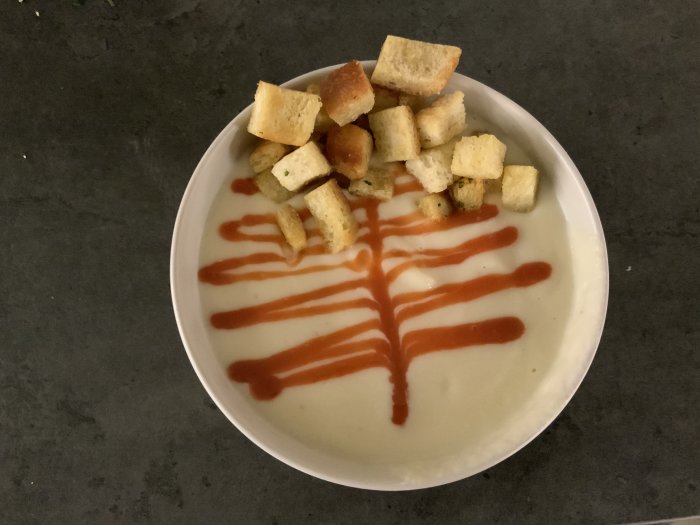 Blomkålssoppa med knaperstekt bacon, vitlökskrutonger och ett mönster av chilisås i en vit skål.