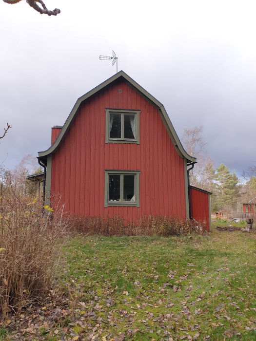 Rödmålat trähus med vita fönsterkarmar och en väderflöjel på taket mot en molnig himmel.