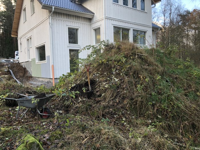 En stor jordhög bredvid vit villa med skottkärra och spade, arbeten för bergvärme pågår.