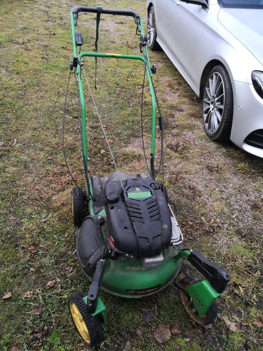 Grön gräsklippare av märket John Deere parkerad på en gräsmatta intill en vit bil.