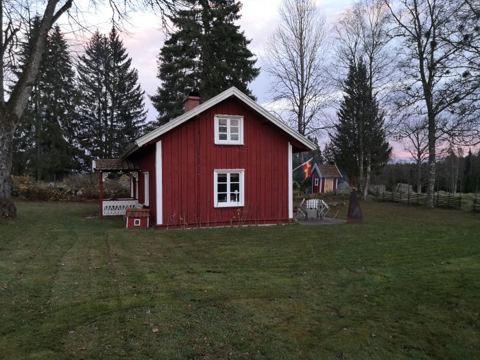 Röd stuga med vita knutar och gräsmatta med spår av mulchning, flaggor och gungställning.