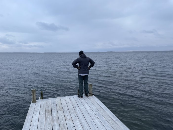 Person står på en träbrygga och blickar ut över ett grått hav under molnig himmel.