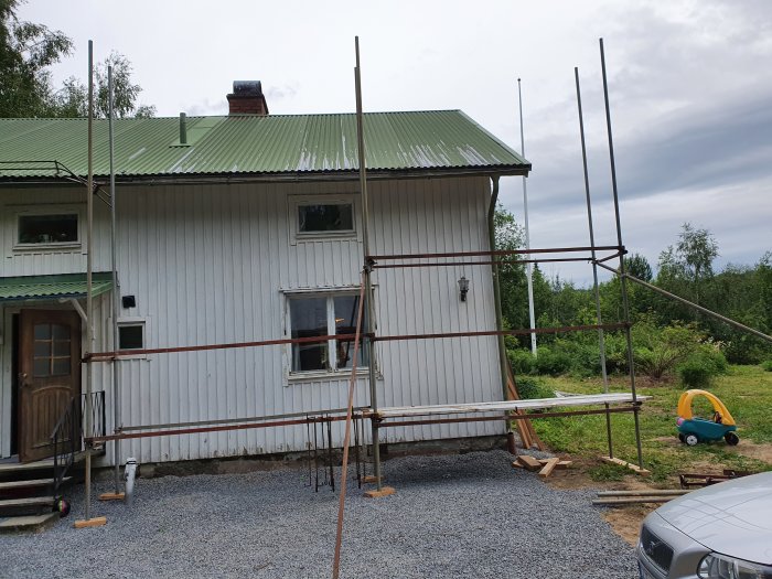 Byggställning av begagnade brädor framför en vit husfasad med grön takplåt och en leksaksbil på gräset.