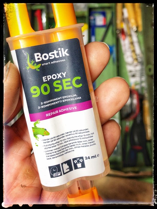 Hand håller en tub Bostik epoxylim med text "EPOXY 90 SEC" i en verkstadsmiljö.