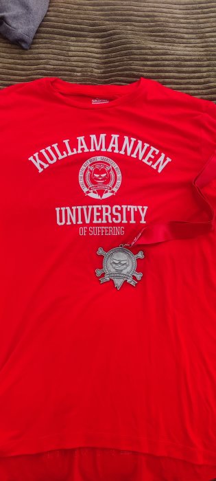 Röd t-shirt med text "Kullamannen University of Suffering", utmärkelse och märke på en mjönstrad bakgrund.