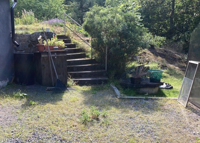 Trädgård med brunnslock till höger om en stentrappa, omgiven av grus och växtlighet.