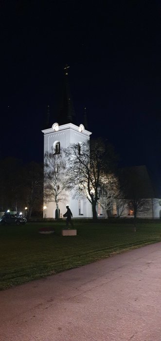 Vit kyrkobyggnad upplyst i natten med ett kors på toppen och en staty framför.