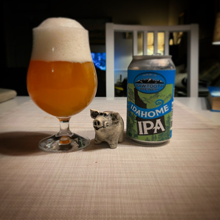 Ett glas öl med tjockt skum bredvid en burk IPA och en liten lergodsgris på ett bord.