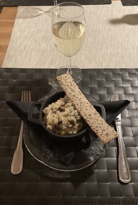 Risotto i gjutjärnsskål med knäckebröd och ett glas vitt vin på dukat bord.