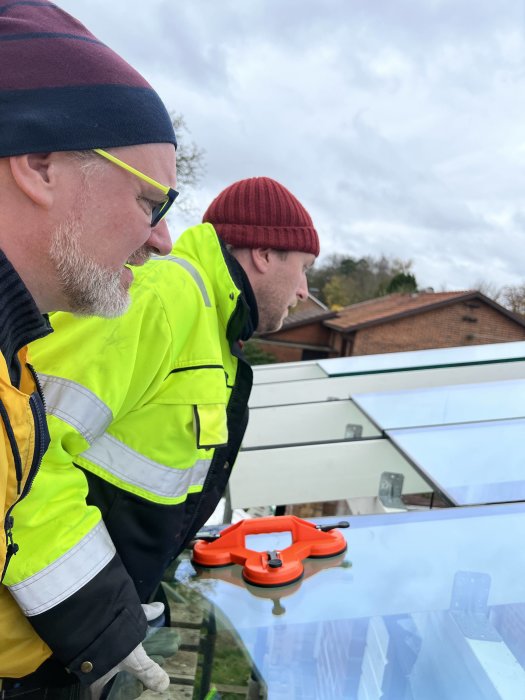 Två personer installerar takglas med hjälp av en sugkoppshållare, med byggnadsställning i bakgrunden.