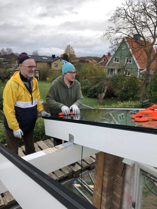 Två personer bär ett stort fönsterglas vid en husbyggnad med ställning och verktyg runtomkring.