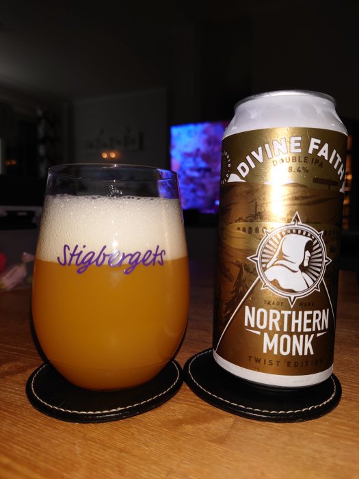 Ölglas fyllt med skummande öl bredvid en burk av Northern Monk Double IPA på ett bord, med suddig bakgrund.