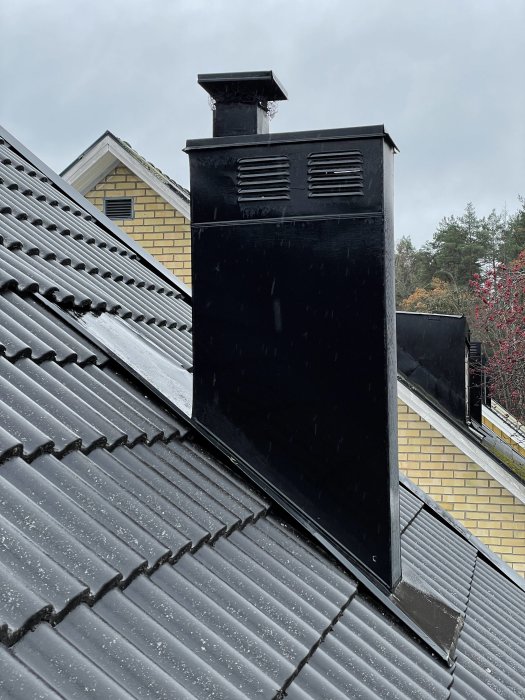 Svart skorsten med ventilation på ett tak med tegelpannor, några regndroppar syns i luften.