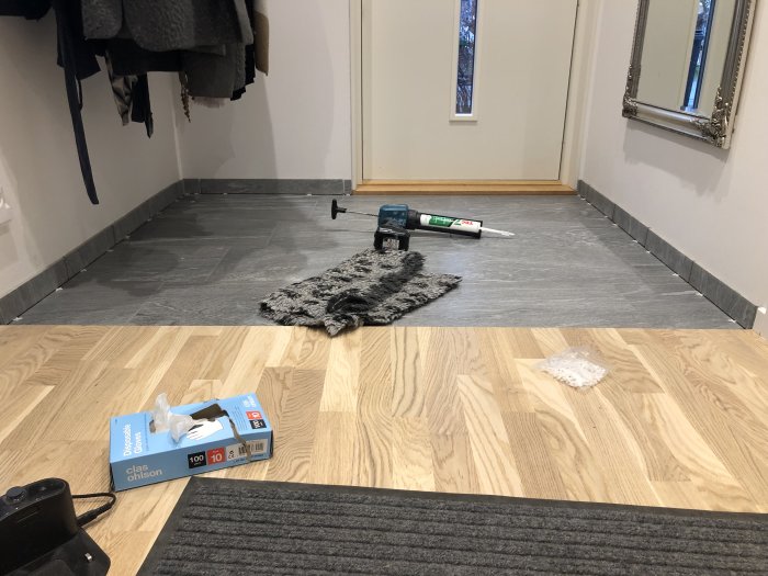 Rum under renovering med limtuber och nya golvsocklar mot grått golv, verktyg och handske på golvet.