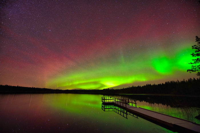 Norrsken över en sjö med en brygga och stjärnklar himmel.