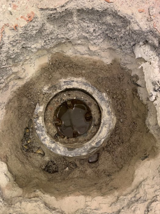 Uppbrytet källargolv med synlig gammal gjutjärnsbrunn och vattenläckage runt kanterna i en tvättstuga under renovering.