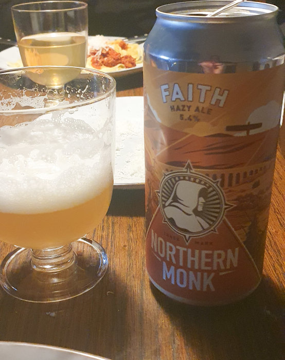 Ölglas med skum, burk märkt "Faith Hazy Ale" och glas med äppeldricka på bord.