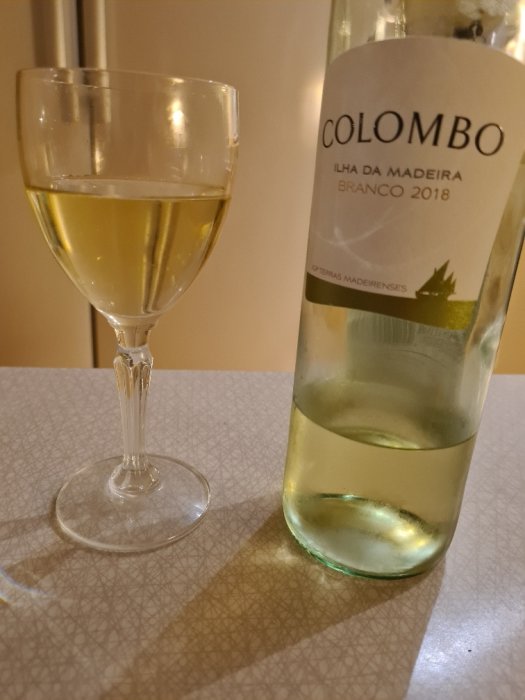 Ett glas vitt vin bredvid en flaska Colombo Madeira Branco 2018 på ett mönstrat bord.