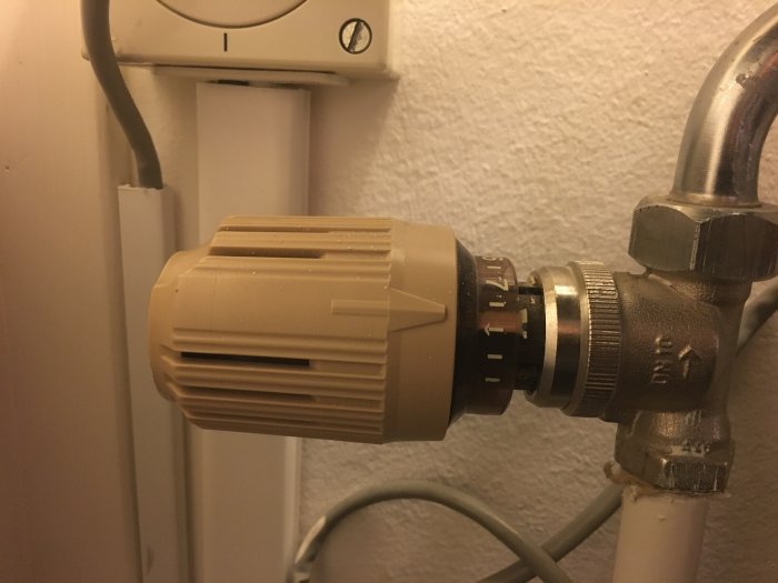 Termostat fastsatt på en radiatorventil bredvid en vit eluttag, termostaten ser ut att vara i behov av att tas bort.