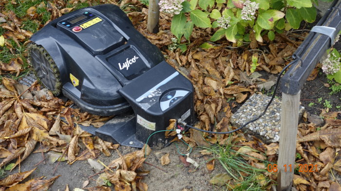 Robogräsklippare vid laddstation med slarvigt lagda kabelskarvar och löv på marken.