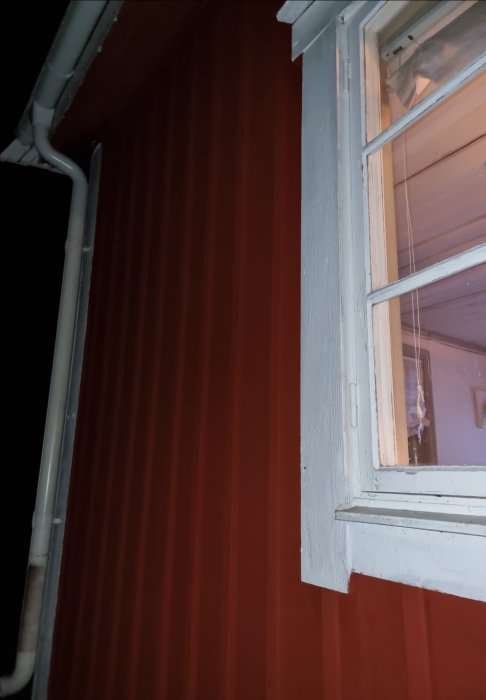 Ett fönster med vit karm på en röd väggbeklädnad vid nattetid; synligt fönsterfoder.