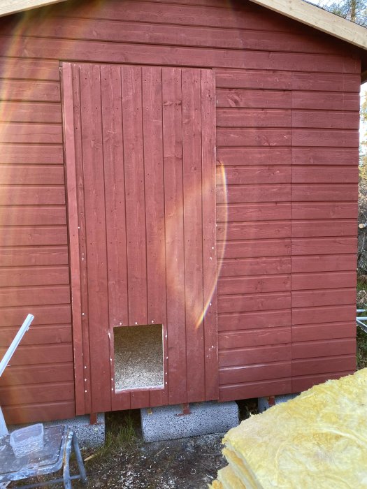 Rödmålat hönshus med öppen liten fyrkantig lucka nära marken, isolering i förgrunden, påbörjade byggnadsarbeten.