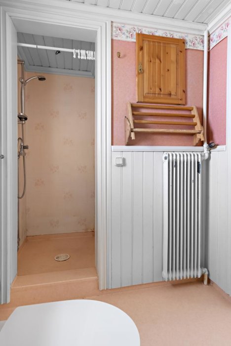 Ett gammaldags badrum med rosa tapet, en duschhörna, vit radiator, och en öppen trätrappa upp till en vindslucka.