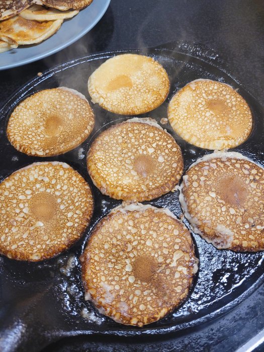 Stekande plättar i en het panna, med en tallrik staplade plättar i bakgrunden.