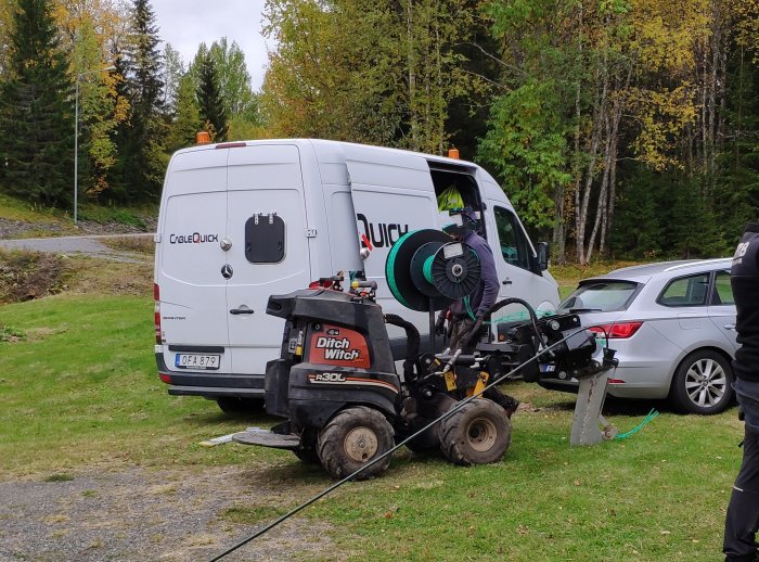 Ditch Witch maskin används på en tomt med en vit skåpbil och person med skyddsutrustning i bakgrunden.