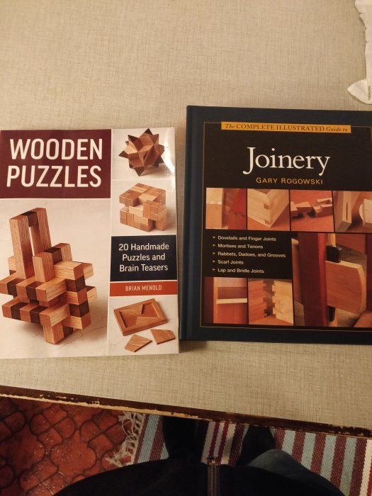 Två böcker om träarbete, "WOODEN PUZZLES" och "The Complete Illustrated Guide to Joinery", på ett bord.