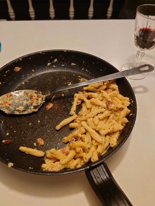Stekpanna med hemlagad carbonara och spatel, ett glas rödvin i bakgrunden på en vit duk.