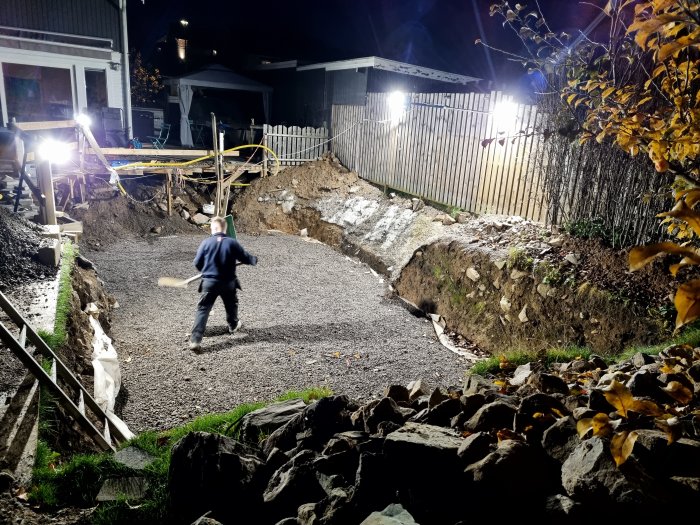 Person jämnar ut grus i en upplyst grävd grund på kvällen vid en byggarbetsplats, omgiven av sten och grävmaskiner.
