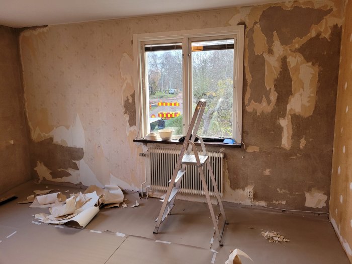 Vägg med delvis borttagen tapet, puts synlig, stege och rivna tapetbitar på golvet.