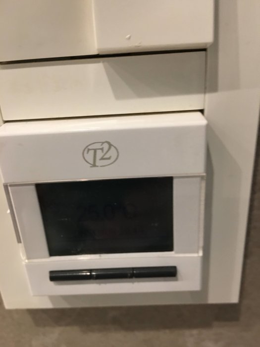 T2 termostat för golvvärme visar låst skärm med texten "26.0C LÅST" på en vit vägg.