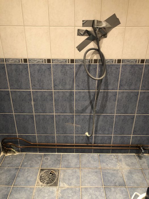 Kaklad badrumsvägg med synliga kopparrör och duschslang fixerad med silvertejp.