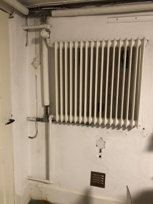 Vit radiator med anslutande rör på en sliten vägg med flagnande färg och ventilkåpa.