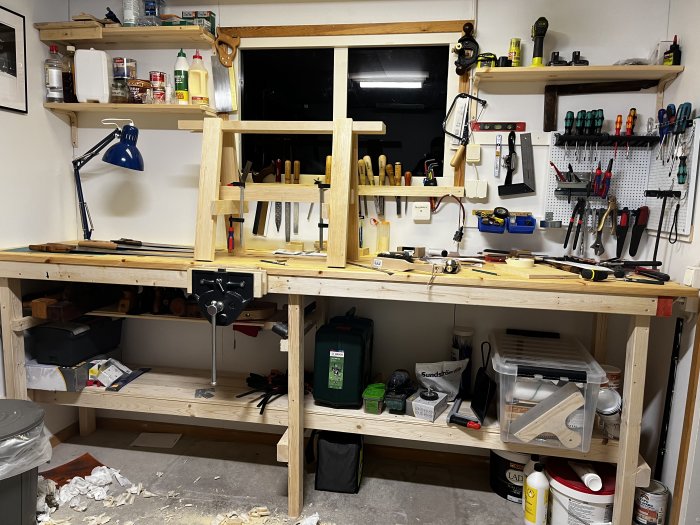 Verktyg och material på en välorganiserad arbetsbänk i ett garage med hyllor och verktygstavlor.