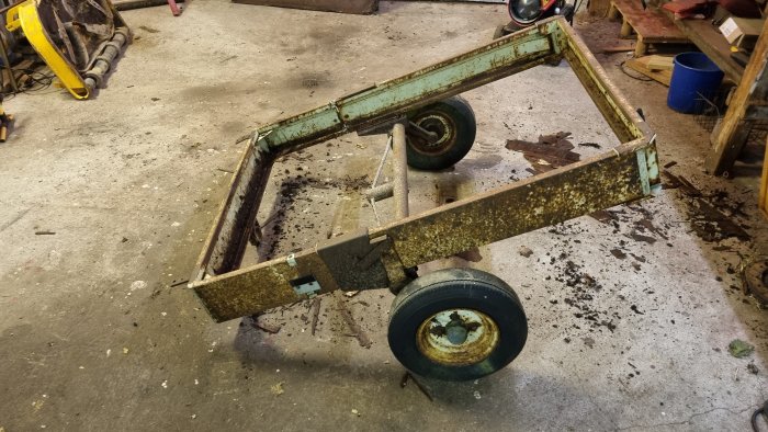 Gammal rostig bilsläp utan botten och dragstång som omvandlats till en parkvagn, stående i ett verkstadsgolv.