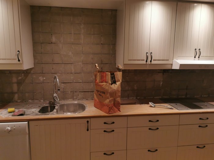 Kök under renovering med borttaget kakel och synlig betongvägg, skåp kvar ovanför arbetsyta.