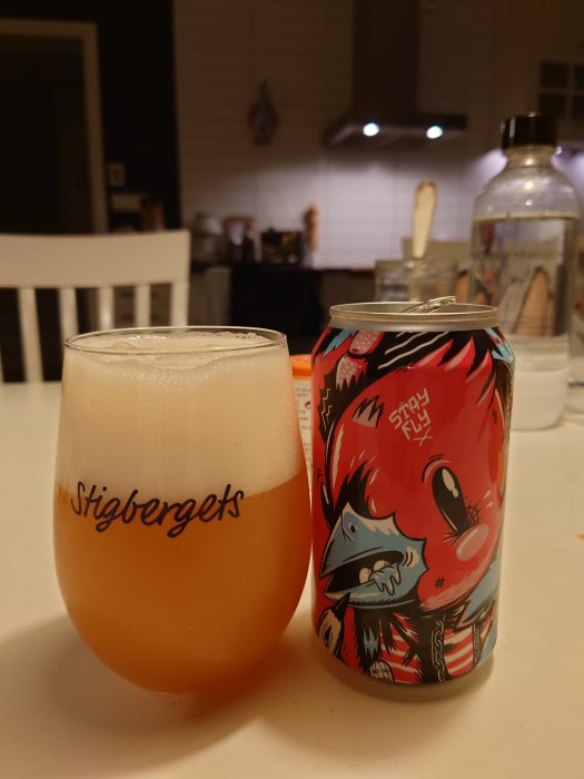 Ett glas fyllt med öl bredvid en färgglad ölburk med konstnärlig design på ett vitt köksbord.