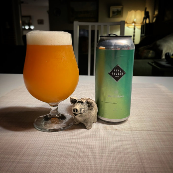 Ölglas med skum krona, ölburk och prydnadsgris på bord.