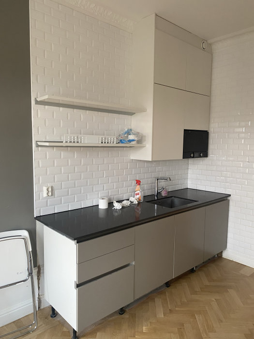 Kök med grå skåpluckor, vit kakelvägg och svart arbetsyta som visar tecken på slarvigt fogat kakel.