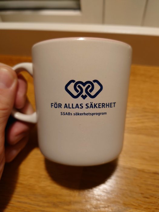 Hand håller en vit mugg med texten "FÖR ALLAS SÄKERHET, SSABs säkerhetsprogram" och grafisk logotyp.